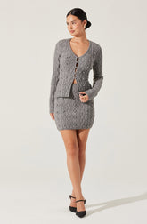 A woman is wearing a matching gray knit cardigan and a fitted gray knit mini skirt, both featuring a diamond cable pattern. The cardigan has a round neckline and buttons down the front, with the top button left open. The skirt is high-waisted, ending above the knees. She is also wearing black pointed-toe heels with ankle straps.